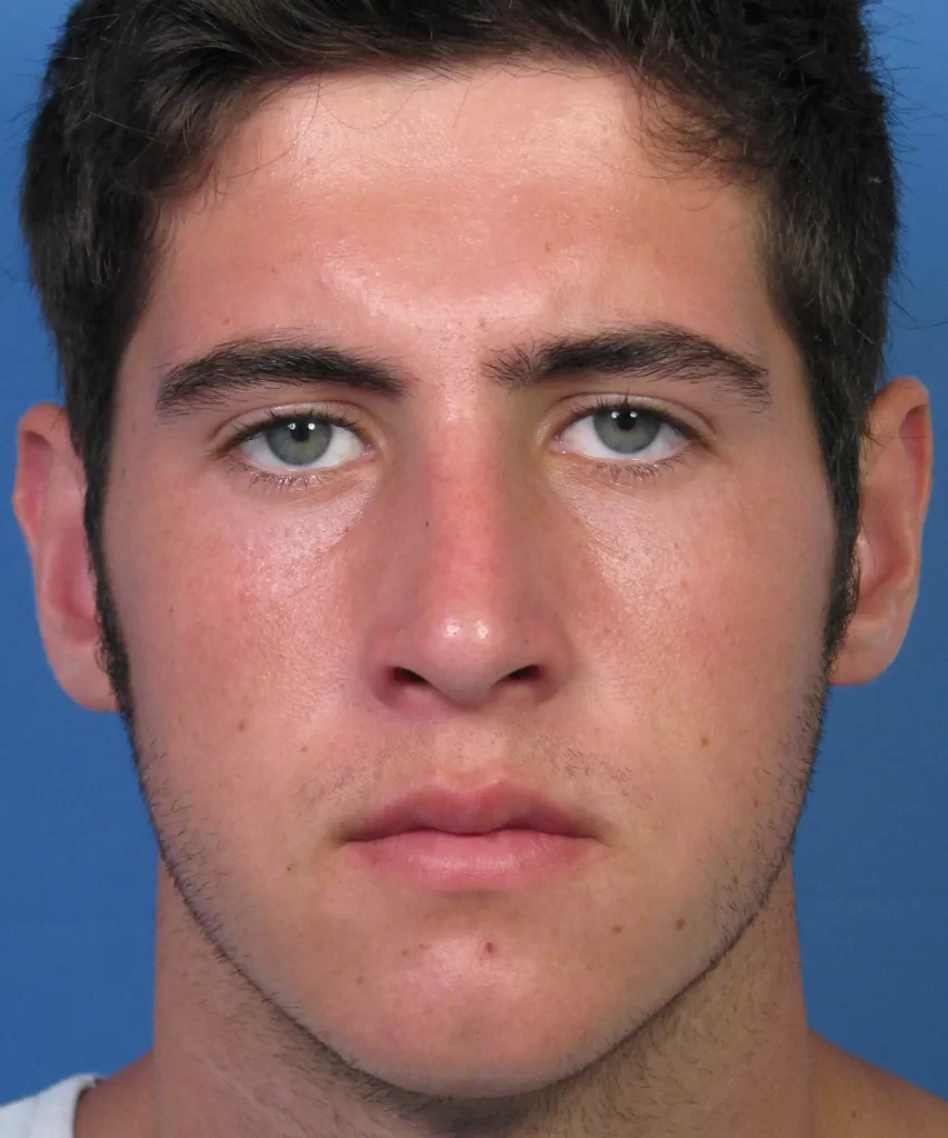 A man with short dark hair and facial stubble, wearing a plain shirt, looks directly at the camera against a blue background.