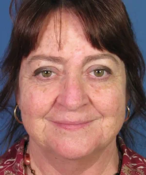 A woman with brown hair and earrings smiles slightly against a blue background. She is wearing a patterned top.