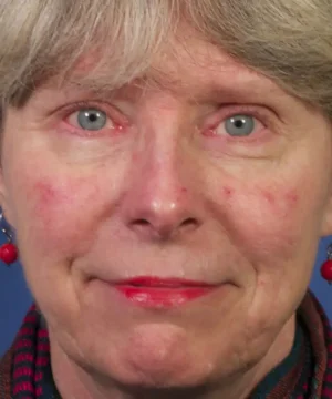 A person with short gray hair and red earrings, wearing minimal makeup, looks directly at the camera.