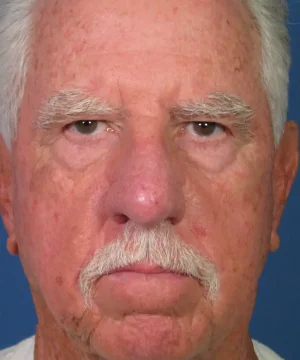 Close-up of an older man with white hair and a mustache, looking directly at the camera against a blue background.