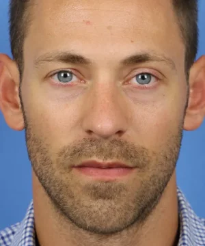 A man with a trimmed beard and short hair is looking directly at the camera against a blue background.