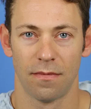 A close-up of a man with short brown hair, blue eyes, and a neutral expression against a blue background.