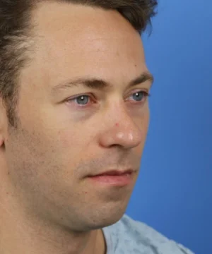 A man with short brown hair and blue eyes, wearing a light blue shirt, looks slightly to the left against a blue background.