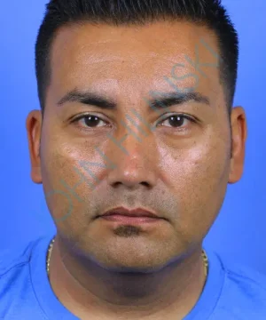 The image shows a man with short dark hair, wearing a blue shirt, against a blue background.