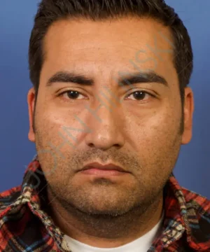 Portrait of a person with short dark hair and beard, wearing a plaid shirt, against a blue background.