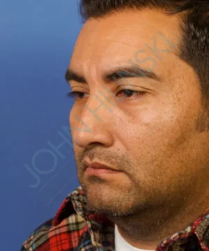 Profile of a man with facial hair wearing a plaid shirt against a blue background.