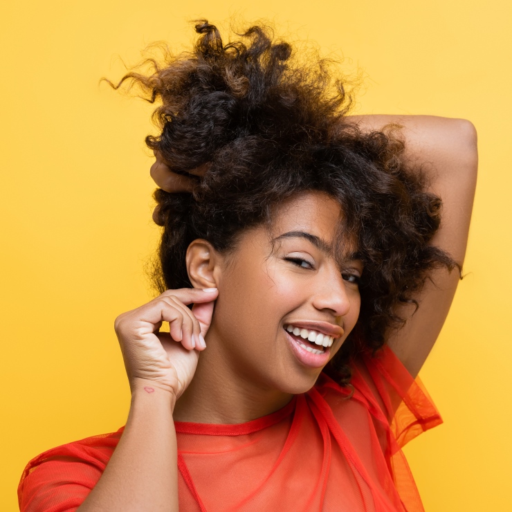 Person with curly hair smiles while adjusting an earring against a solid yellow background. Wearing a sheer red top.