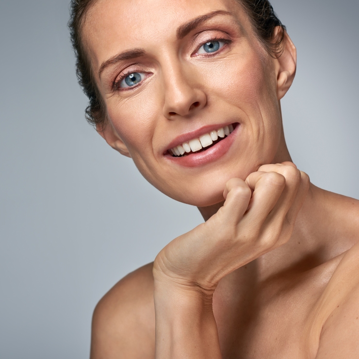 A person with light skin smiles while resting their chin on their hand against a plain background.