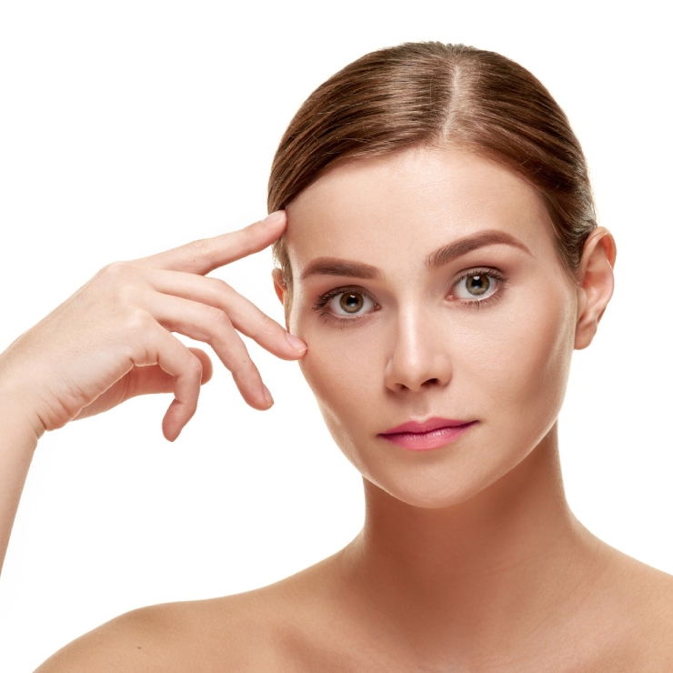 A person with light skin and brown hair is posing against a white background, touching their temple with their right index finger.