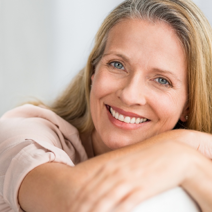 Smiling woman with long blonde hair, resting her arms on a surface, wearing a light-colored blouse.