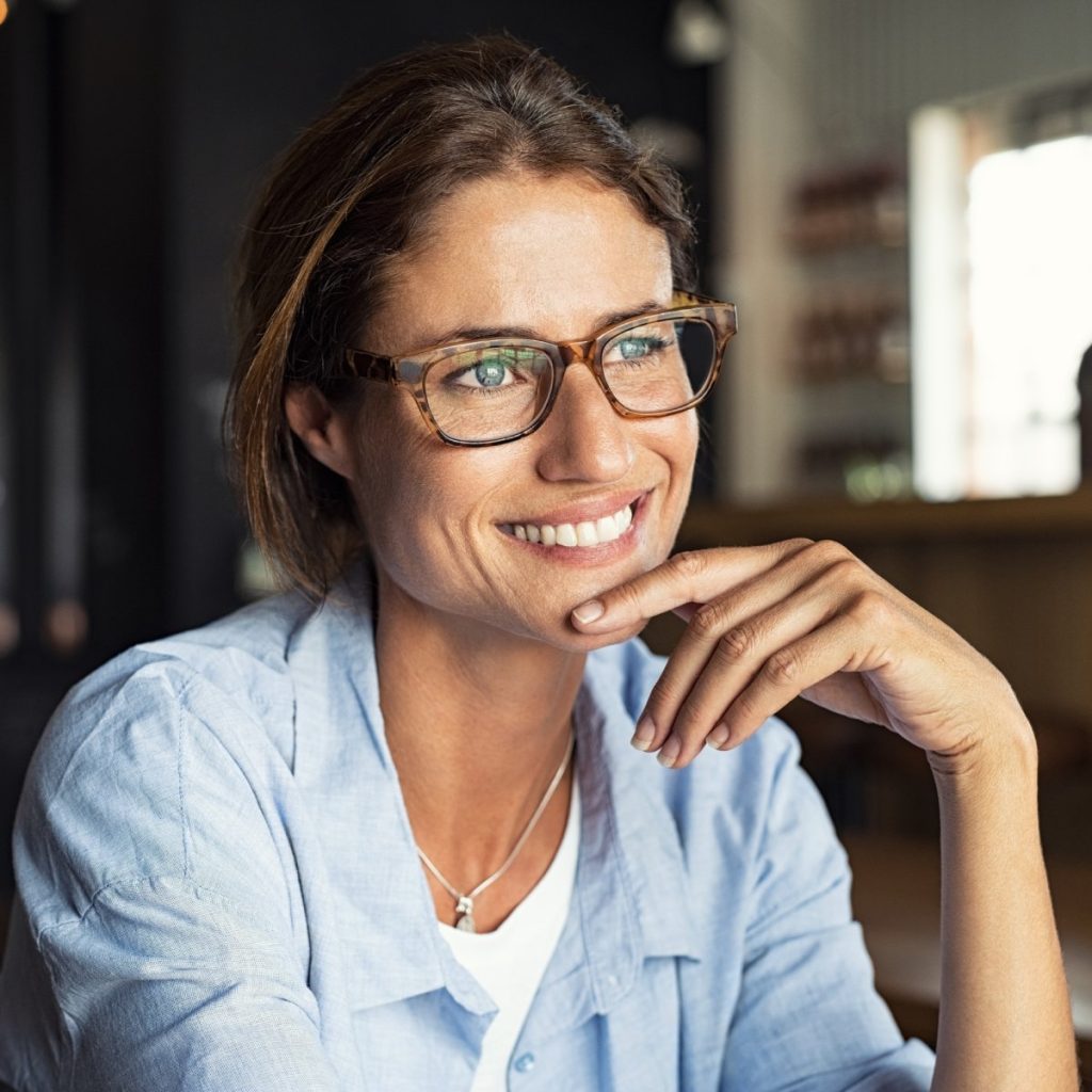 A woman wearing glasses and a light blue shirt smiles while resting her chin on her hand in a softly lit indoor setting.