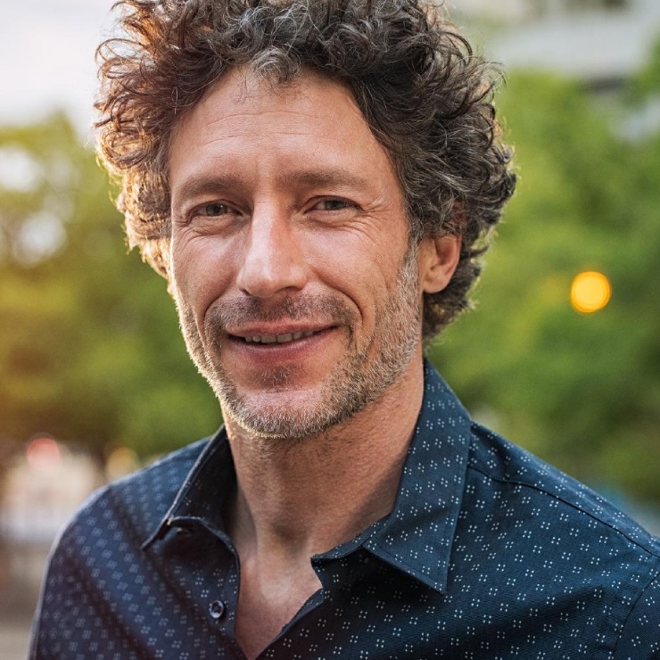 A man with curly hair and a slight beard smiles outdoors, wearing a dark patterned shirt. Trees and blurred lights are in the background.