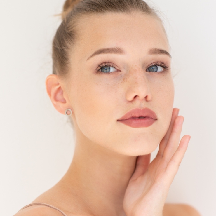 A person with light hair pulled back, light eyes, and freckled skin is touching their face gently, looking directly at the camera with a neutral expression.