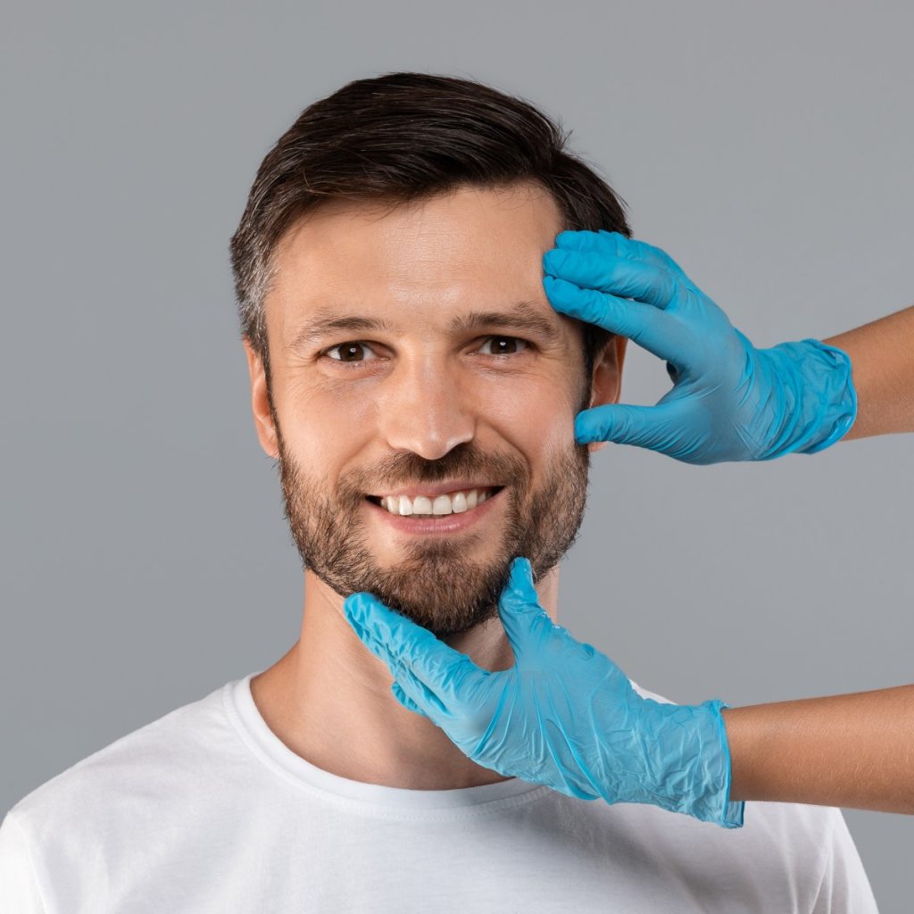 A man smiling while a person in blue gloves touches his face against a gray background.