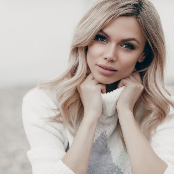 A woman with long blonde hair wearing a white sweater looks directly at the camera, hands gently holding her collar.