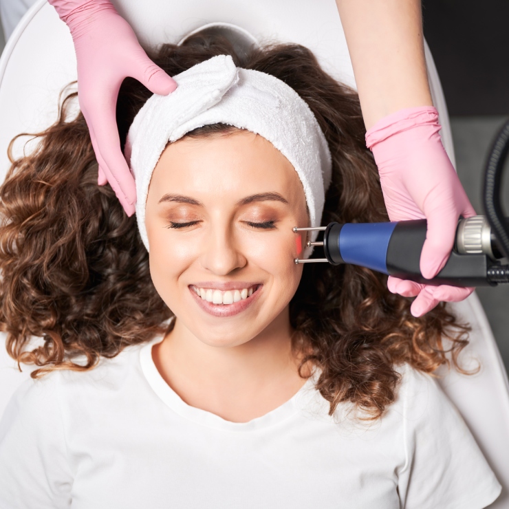 A woman with curly hair receives a facial treatment from a machine, while wearing a white headband. A person in pink gloves holds the equipment. She is smiling and lying on a white surface.