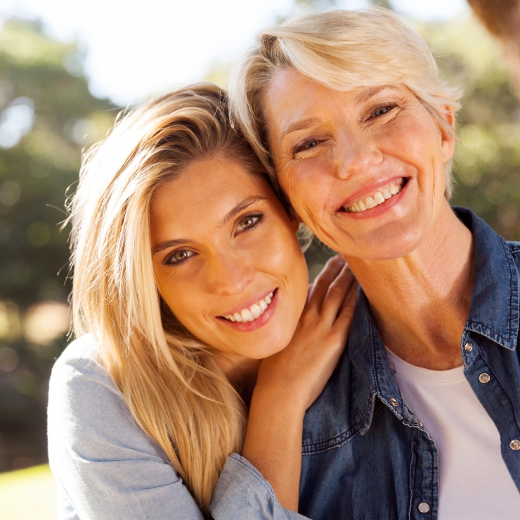 Two people smiling, with the younger person resting their head on the older person's shoulder, outdoors in a sunny setting.