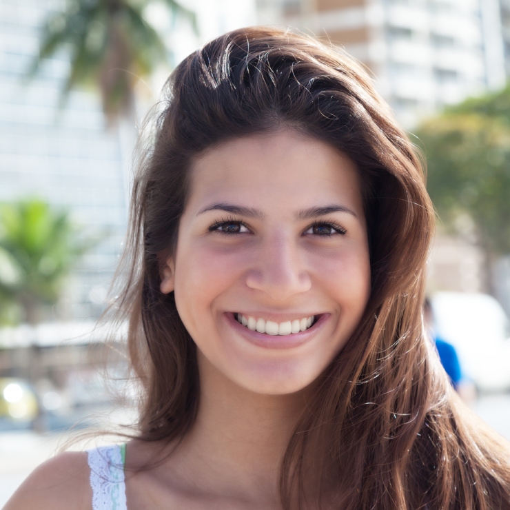 A person with long brown hair smiling outdoors, with palm trees and buildings in the background.