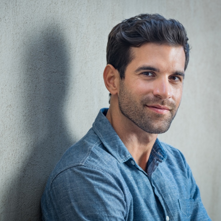 A man with short hair and a beard smiles while leaning against a gray textured wall. He is wearing a blue button-up shirt.