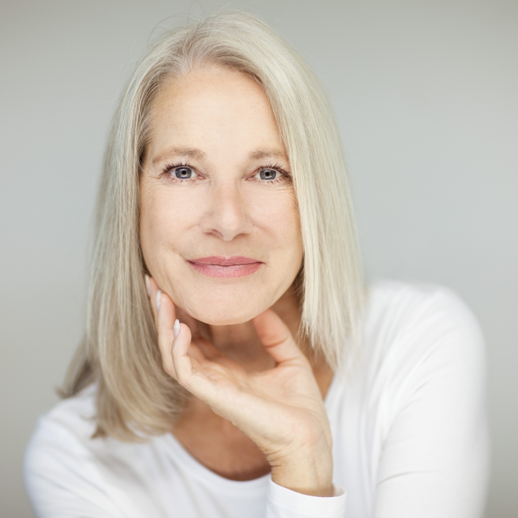 A woman with long, light gray hair and a white top rests her chin on her hand, smiling softly.