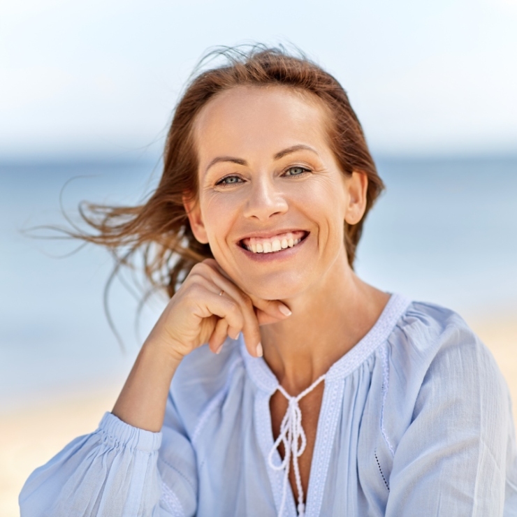 A woman with long hair smiles at the camera, sitting on a beach. She is wearing a light blue blouse. The background features a blurred ocean and sky.