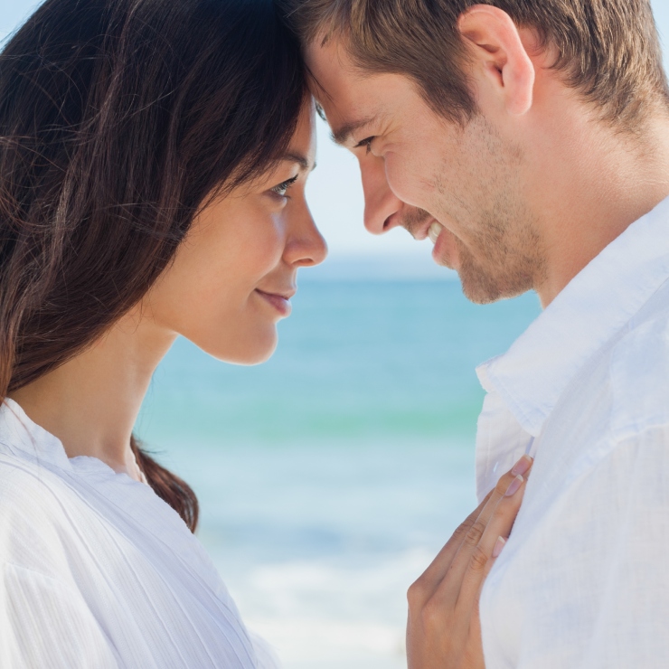 A man and woman stand closely facing each other on a beach, smiling with their foreheads nearly touching.