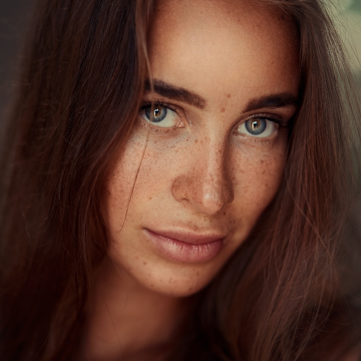 A woman with long brown hair and blue eyes looks directly at the camera. She has freckles and a neutral expression.