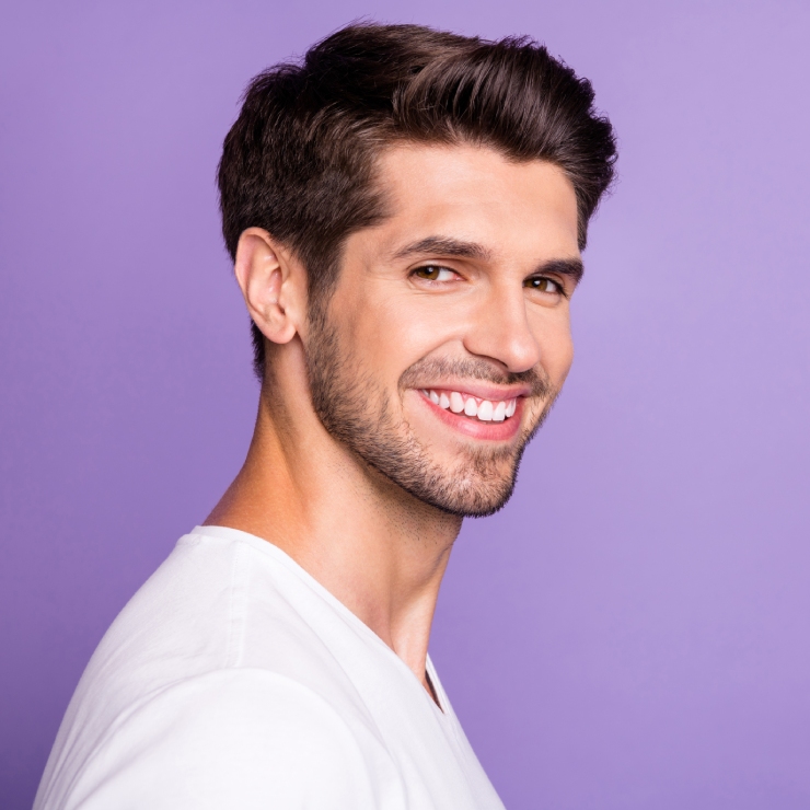 A man with dark hair and a beard smiles, wearing a white shirt against a purple background.