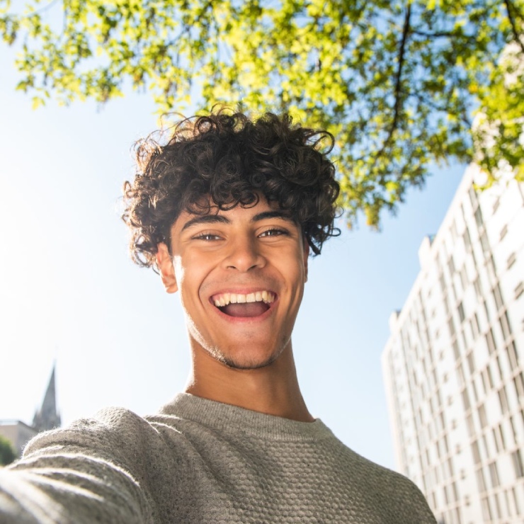 A person with curly hair smiles widely under a tree with sunlight filtering through. A multi-story building and a church steeple are visible in the background.