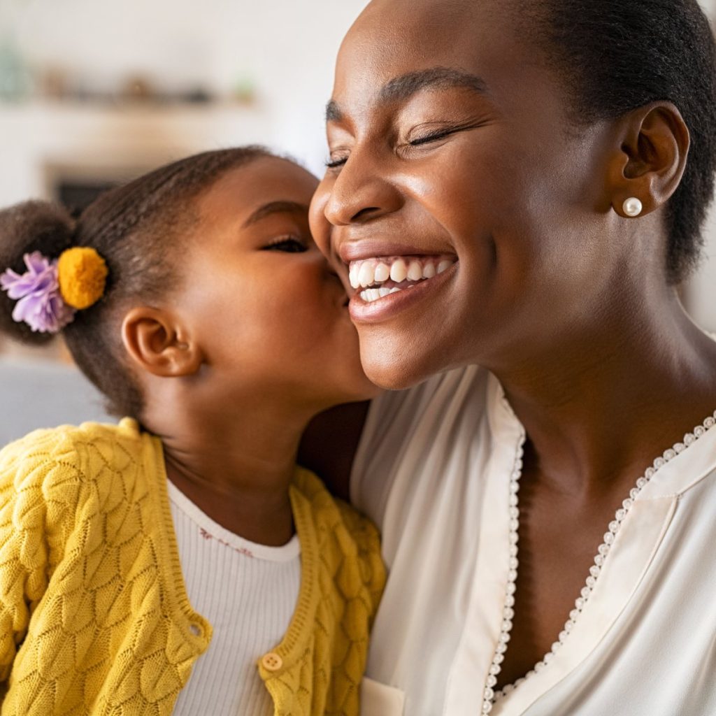 A child kisses an adult on the cheek. Both are smiling warmly. The child wears a yellow sweater and the adult wears a white blouse.