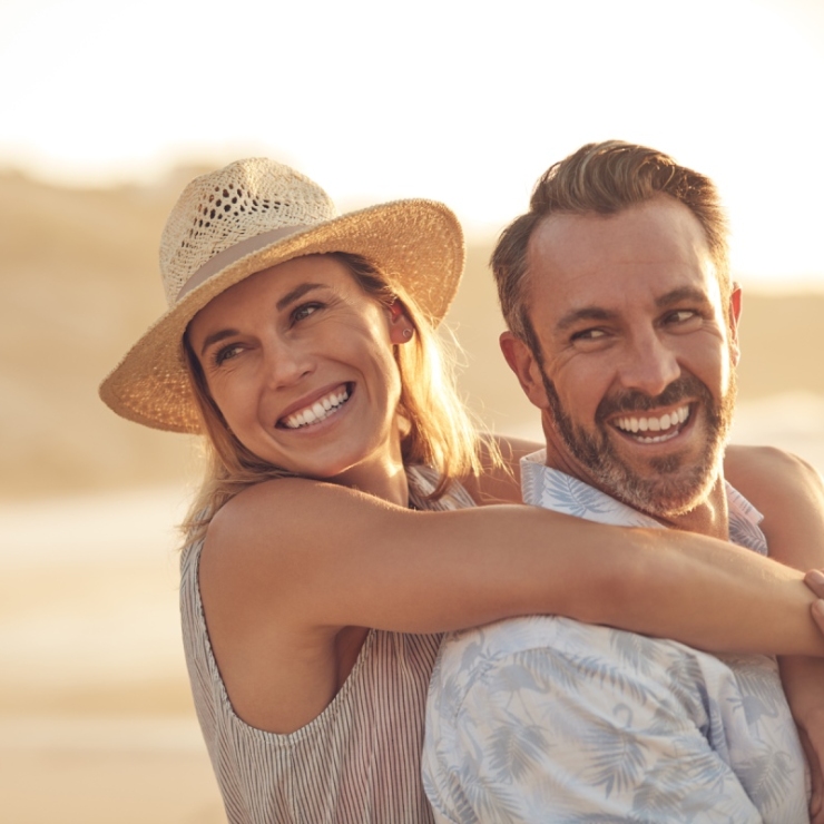 A woman in a straw hat smiles and hugs a man from behind. They are both smiling and standing on a sunny beach.