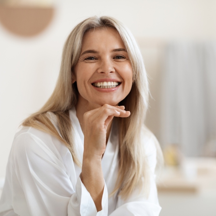 A woman with long blonde hair smiles while sitting in a bright room, wearing a white top.