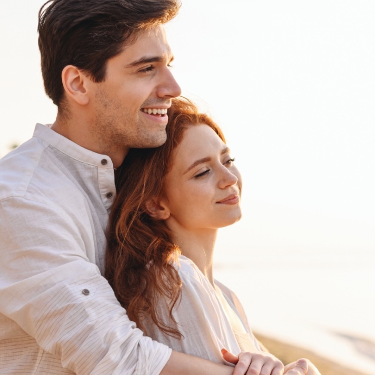 A man and a woman smile while standing on a beach at sunset. The man is behind the woman, holding her with both arms.