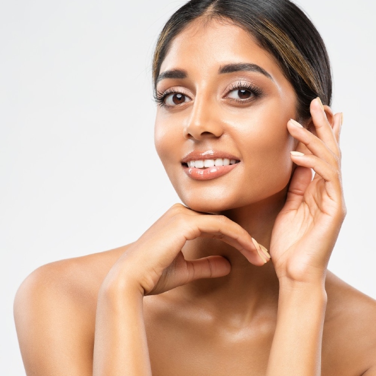 Woman with a clear complexion smiles while posing with one hand near her face and the other under her chin against a plain background.