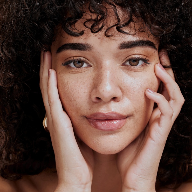 Person with curly hair and freckles gently touching their face with both hands.