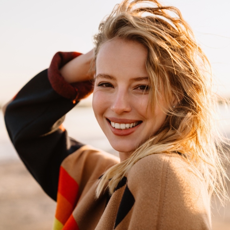 Person smiling at the camera, with wind-swept hair, wearing a colorful coat, outdoors on a sunny day.