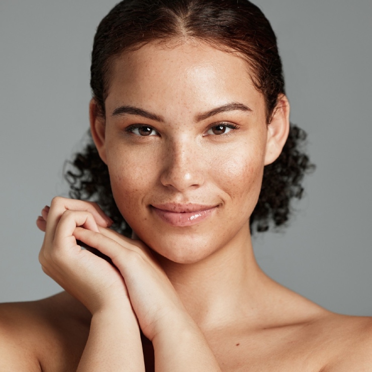 Portrait of a young woman with curly hair smiling softly, hands gently touching her cheek, against a plain gray background.