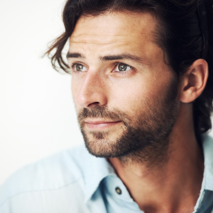 A man with brown hair and a beard gazes to the side. He is wearing a light blue shirt.