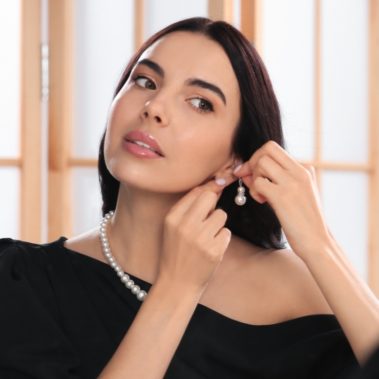 A woman in a black dress adjusts a pearl earring while wearing a matching pearl necklace.