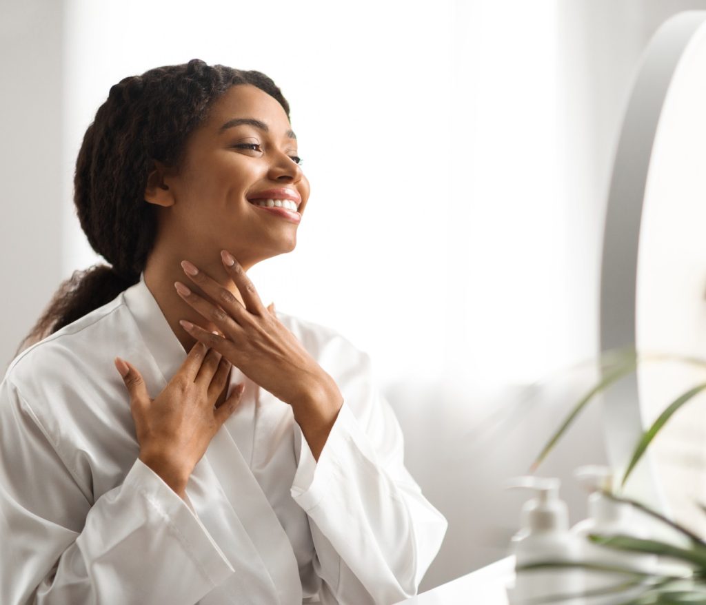 A woman in a white robe smiles while touching her neck, looking at herself in the mirror.