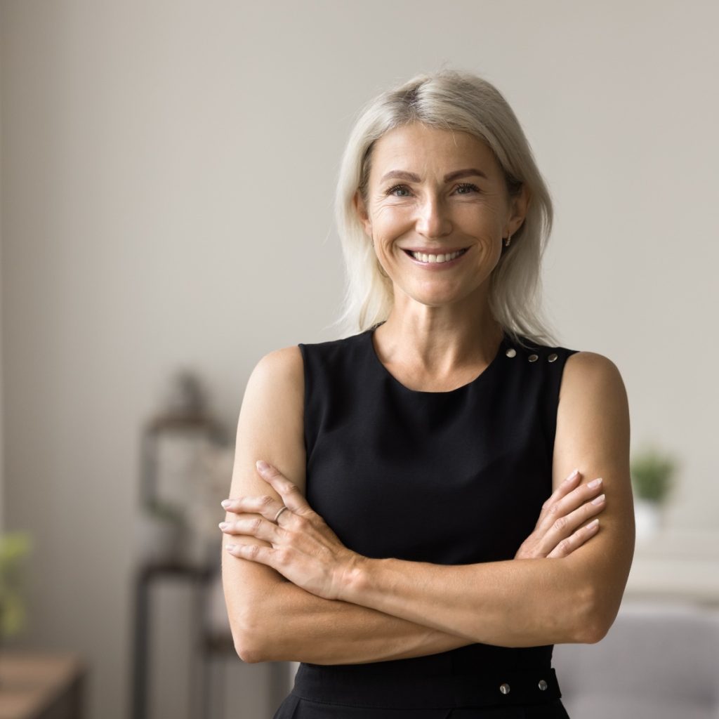 A smiling woman with light hair stands with arms crossed, wearing a sleeveless black top, in a softly lit room.