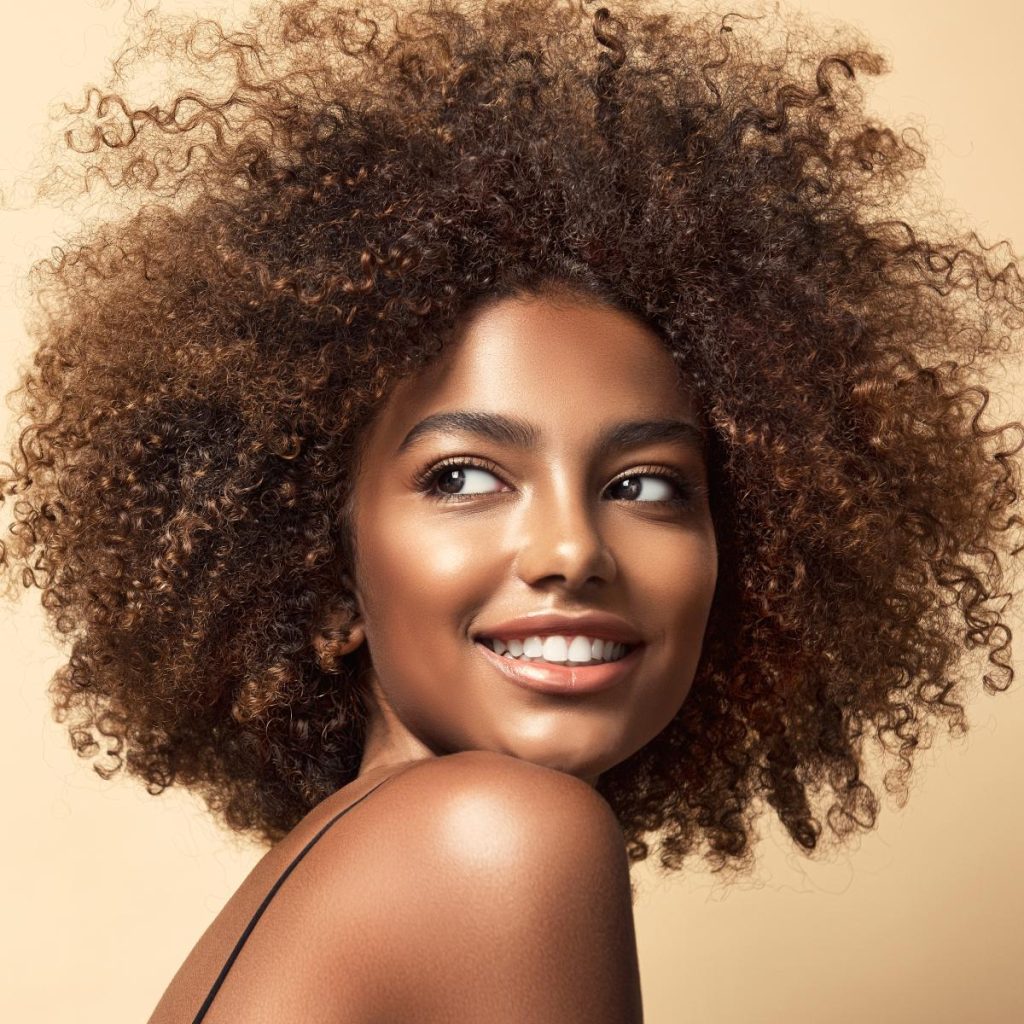 A person with voluminous curly hair smiles while looking to the side, set against a plain beige background.