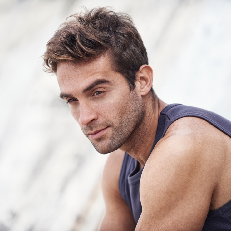 Man with short brown hair wearing a sleeveless shirt, looking off to the side against a blurred background.