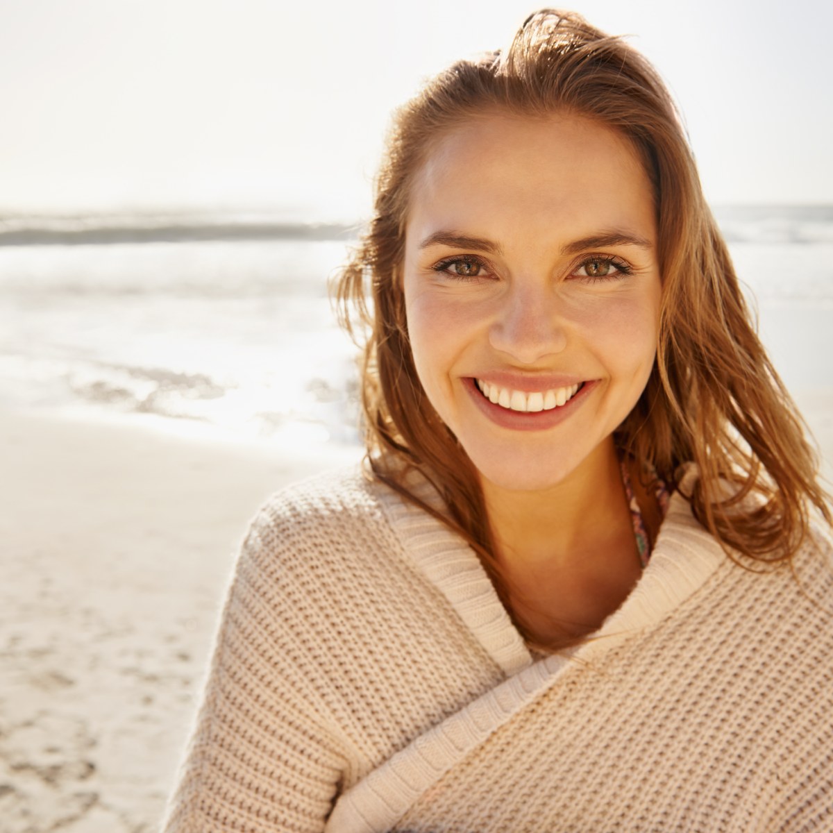 Smiling person standing on a sunny beach, wearing a beige sweater.