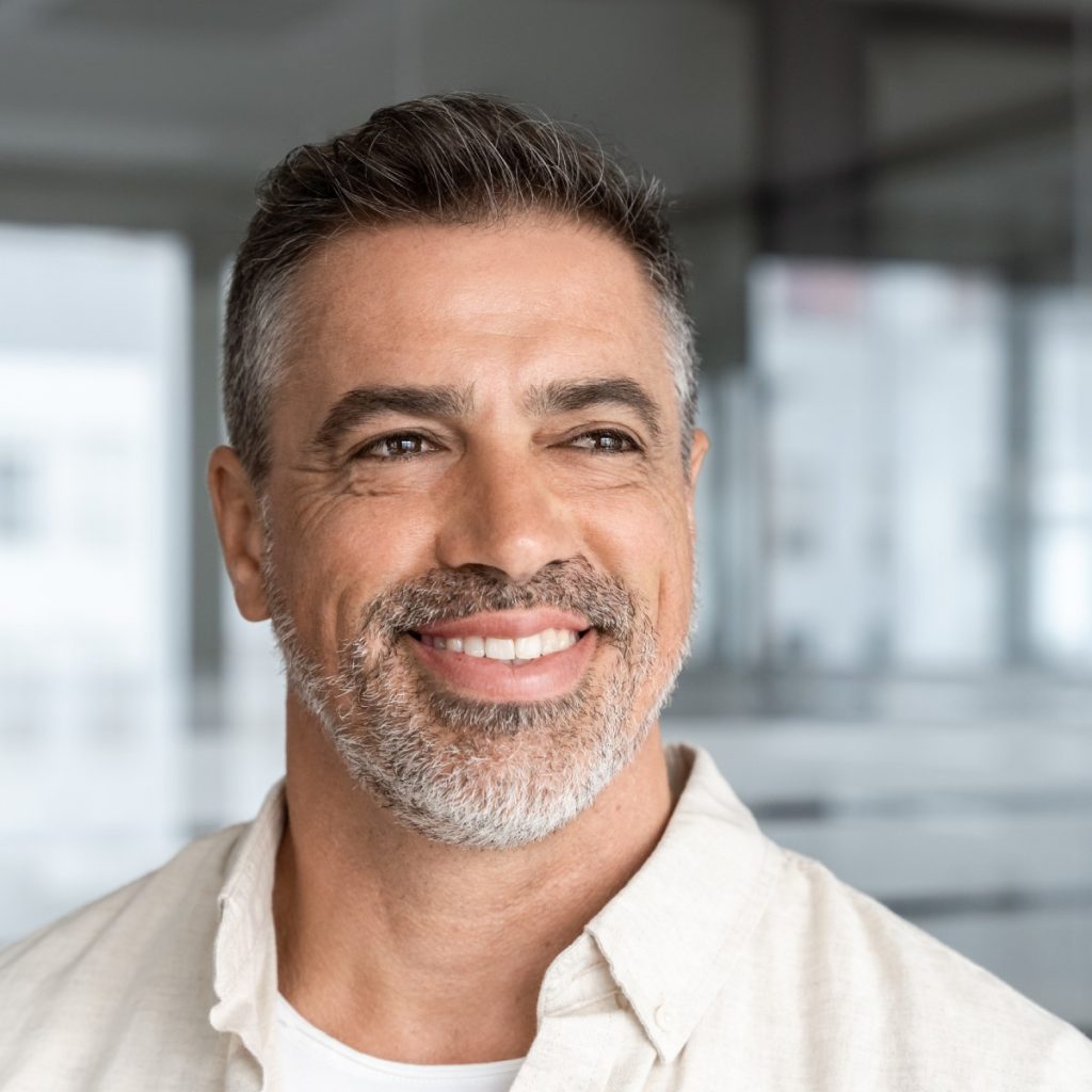 A man with short gray hair and a beard smiles while looking away from the camera in a soft-lit room.