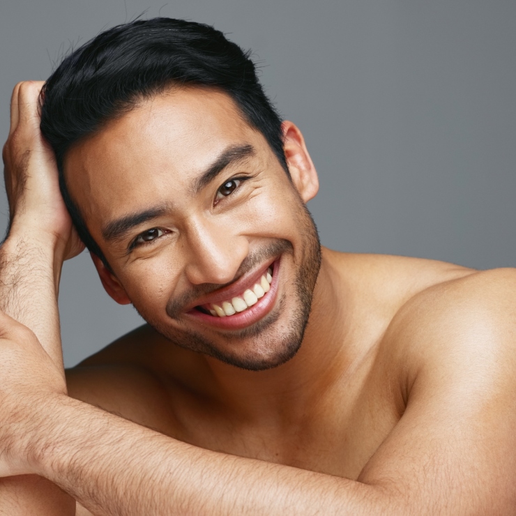 A man with dark hair and a beard smiles while resting his head on his hand against a gray background.