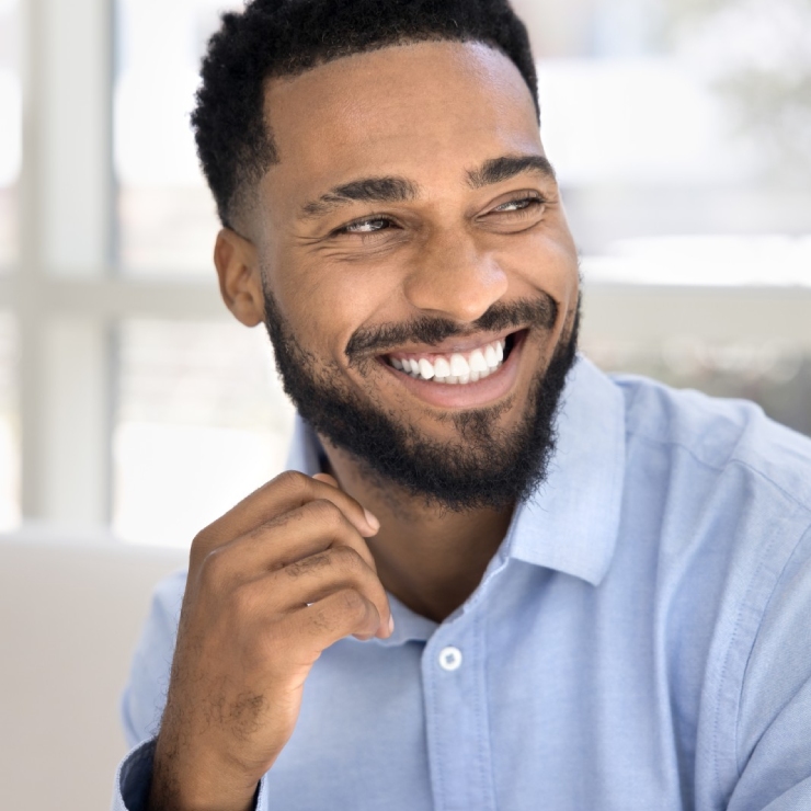 A man with a beard smiles while wearing a light blue shirt. The background is bright and blurred.