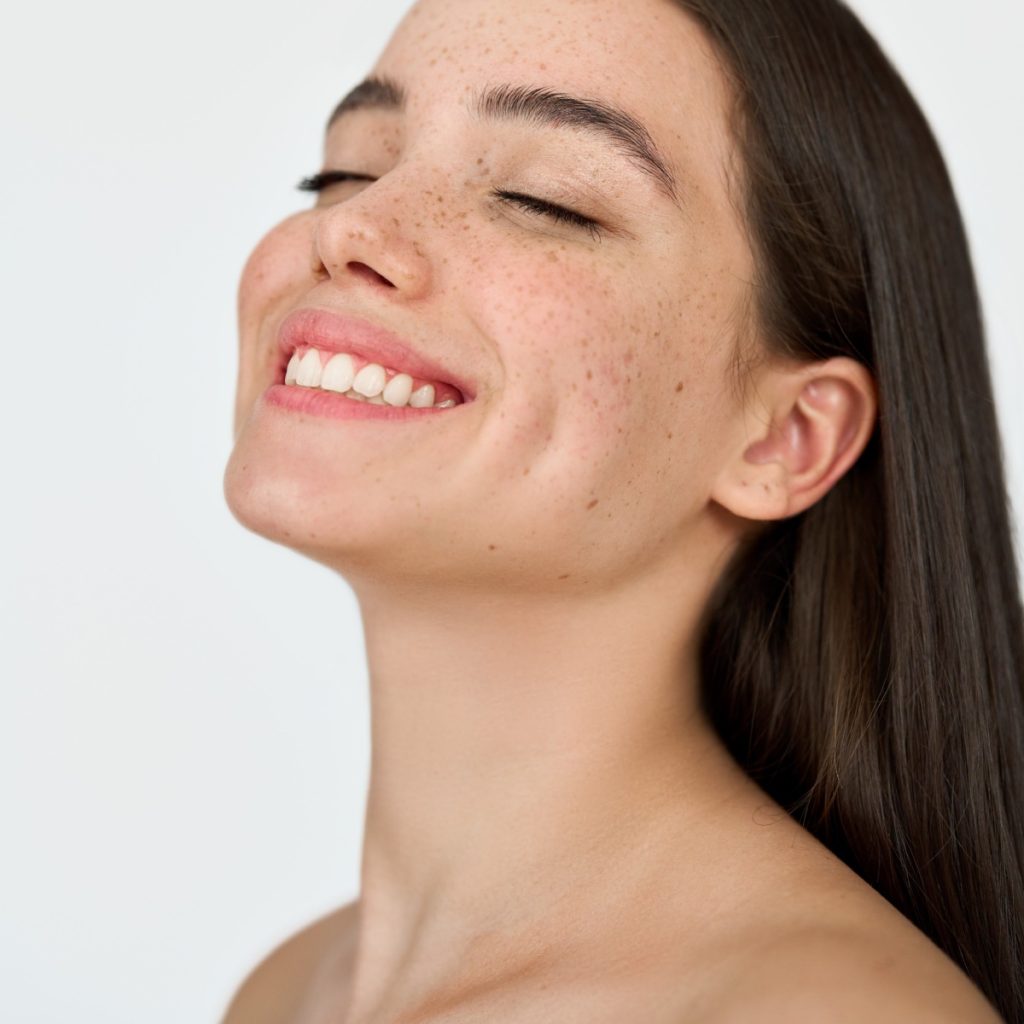 A person with long, dark hair and freckles smiles with eyes closed, head tilted back slightly, on a plain white background.