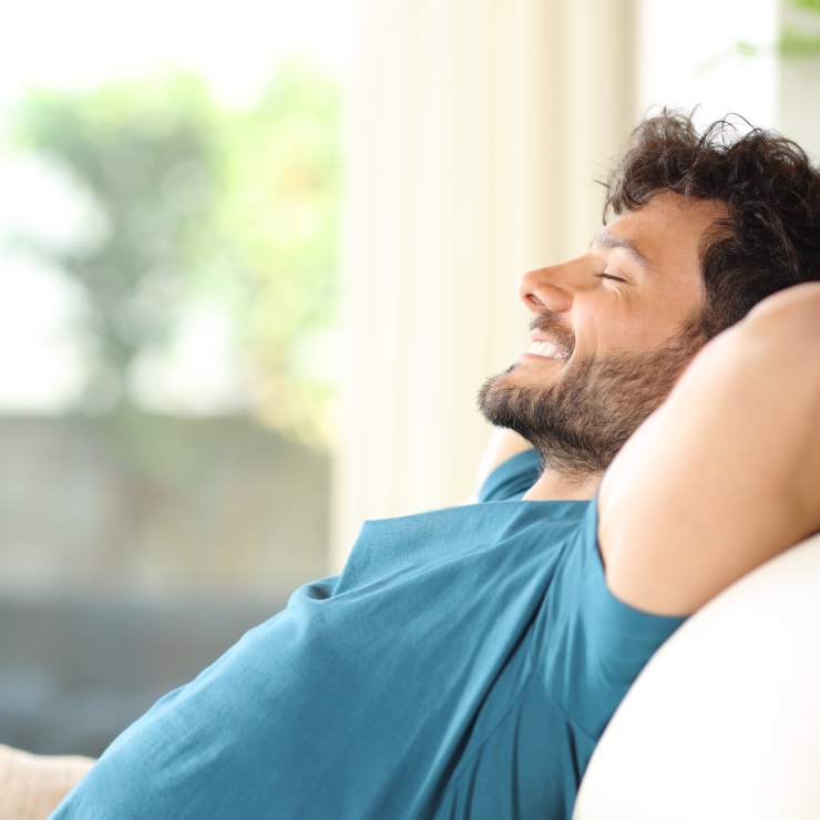 A man with a beard is smiling and leaning back on a sofa, wearing a blue shirt.