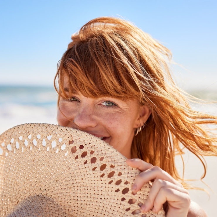 Person with red hair partially hiding their face with a straw hat on a sunny beach.